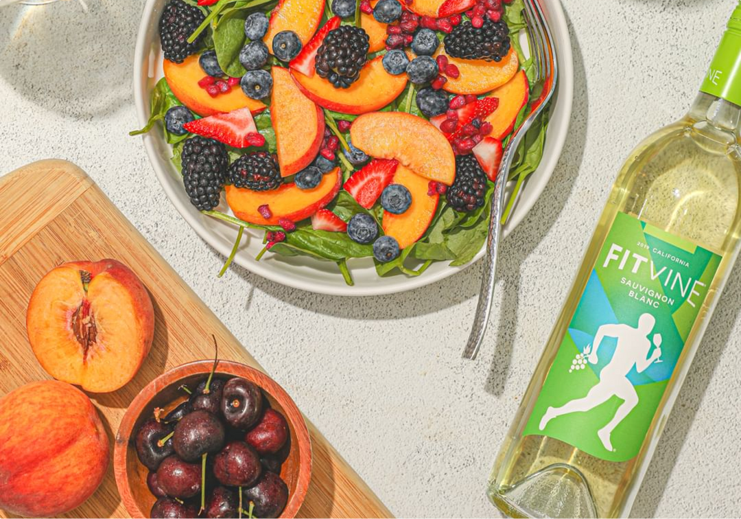 Salad containing blueberries, blackberries, peaches, and spinach next to a bottle of Fitvine's original Sauvignon Blanc
